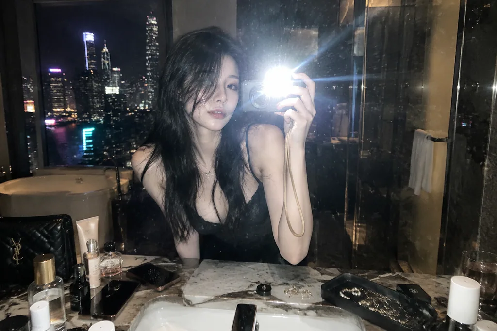 Late-night mirror selfie of a woman in a high-rise marble bathroom over a city skyline
